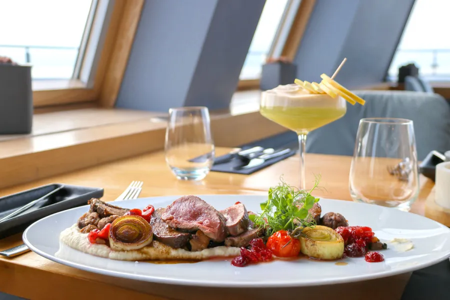 Hauptgericht und Cocktail auf einem Tisch mit großem Fenster und Blick über die Ostsee im Restaurant Weitblick.