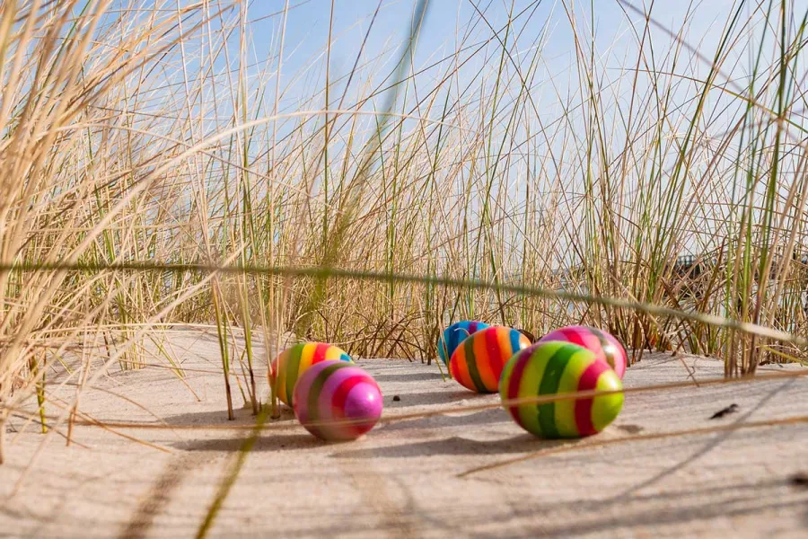 Sechs regenbogenfarben gestreifte Ostereier im Dünensand unter Strandhafer und blauem Himmel, Osterangebot THE GRAND Ahrenshoop