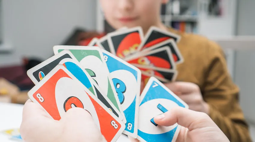 Nachaufnahme von zwei Händen, die UNO-Karten hält mit einem Kind im Hintergrund, der auch UNO-Karten in der Hand hält.