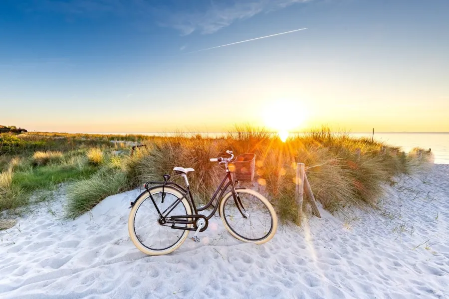 Ein schwarzes Fahrrad steht im Sand vor Dünen am Strand, während die Sonne am Horizont untergeht und warmes Licht über Meer und Gras wirft.