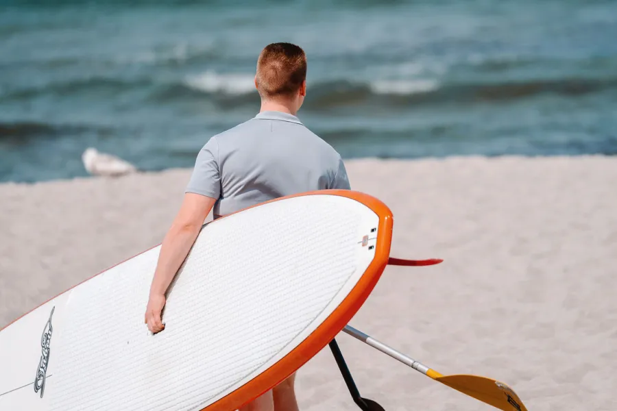 Mann trägt ein Stand-up-Paddle-Board am Strand in Ahrenshoop Richtung Ostsee.