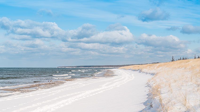 Verschneiter Ostseestrand mit Dünen und sanften Wellen unter blauem Himmel mit Wolken.