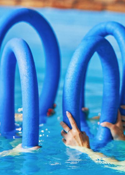 Vier Frauen trainieren beim Aquafitness im Pool mit blauen Schwimmnudeln.