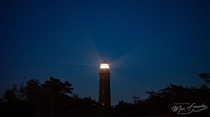 Leuchtturm bei Nacht, sein Lichtkegel strahlt weit in den sternklaren Himmel.