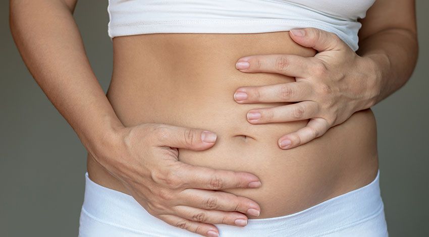 Eine Frau hält sich ihren aufgeblähten Bauch und trägt ein weißes Oberteil sowie eine weiße Hose.