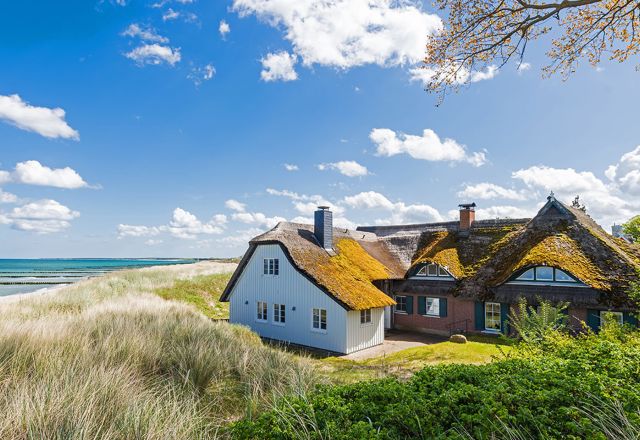 Reetdachhaus an der Ostseeküste mit Blick auf Strand, Dünen und das türkis schimmernde Meer bei Sonnenschein.