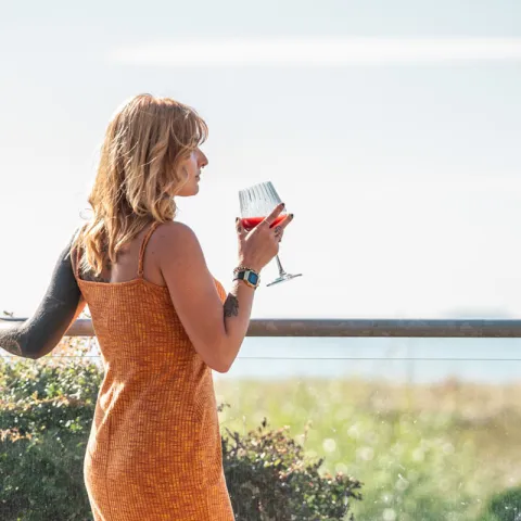 Frau steht auf einer Terrasse mit Drink und Blick auf die Dünenlandschaft.