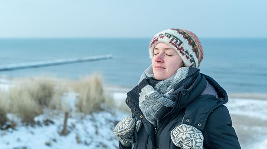 Frau mit Winterkleidung genießt mit geschlossenen Augen die frische Ostseeluft am verschneiten Strand.