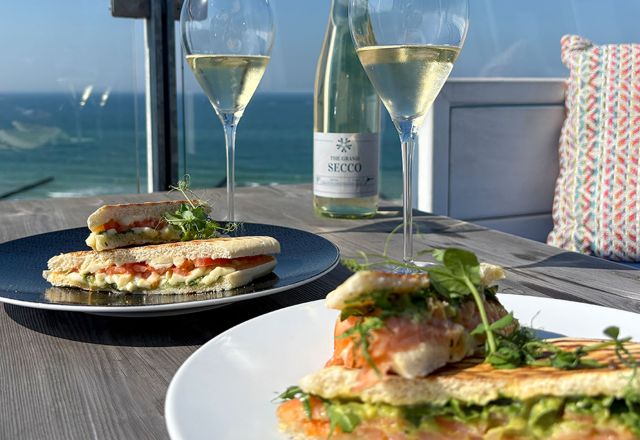 Zwei belegte Fischbrötchen mit Fisch, Salat und Kräutern auf Tellern sowie zwei Gläser Secco auf einem Holztisch mit Blick auf die Ostsee.