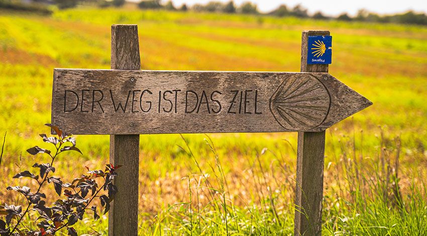 Holzschild mit der Aufschrift „Der Weg ist das Ziel“ auf einer Wiese, daneben das Symbol des Jakobswegs.