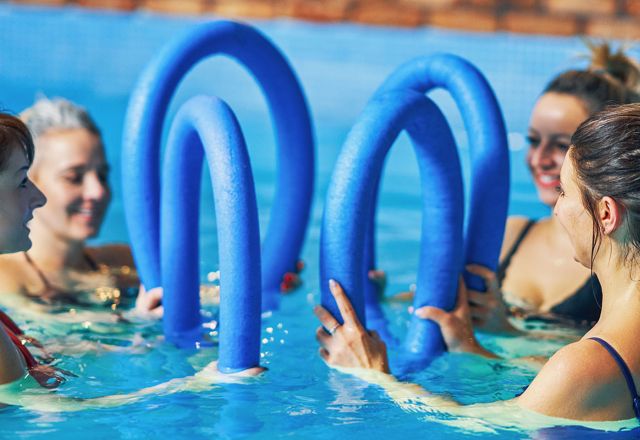Vier Frauen trainieren beim Aquafitness im Pool mit blauen Schwimmnudeln.