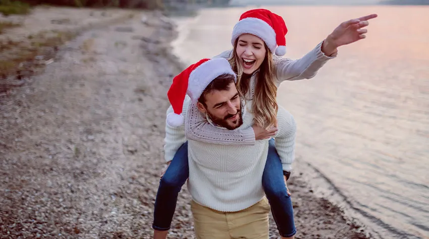 Eine Frau wird huckepack von ihrem Freund getragen. Lachend laufen Sie am Strand entlang und tragen Weihnachtsmützen.