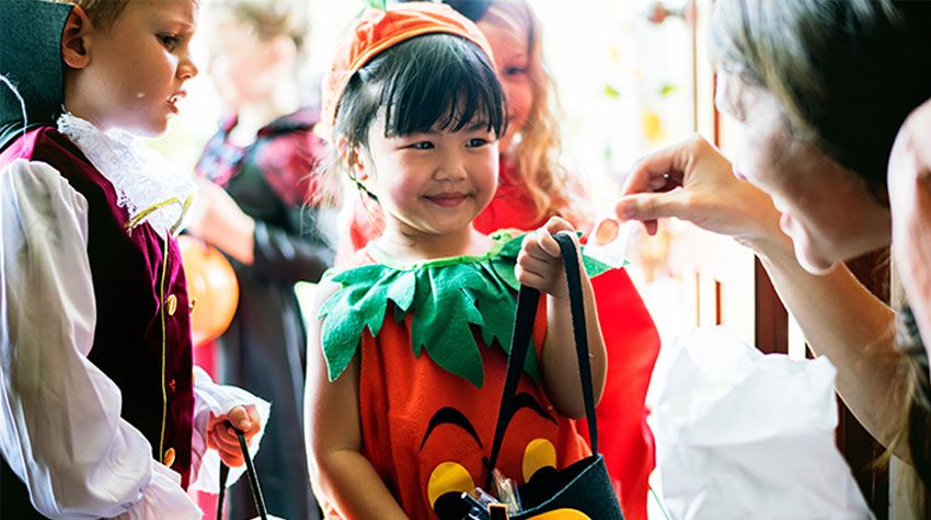 Kinder in bunten Halloweenkostümen gehen von Tür zu Tür und bekommen lachend Süßigkeiten geschenkt.