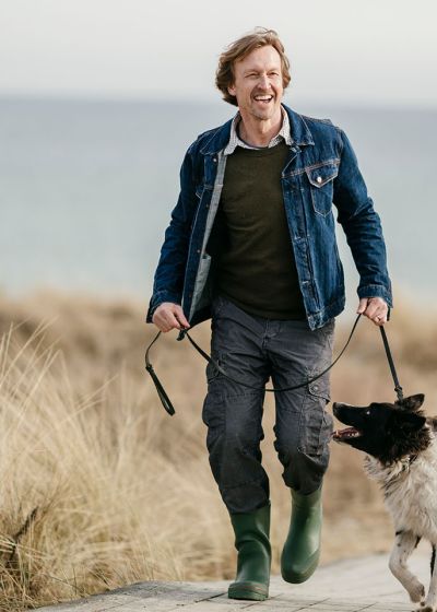 Mann mit Hund an der Leine spaziert entlang eines Strandes. Im Hintergrund Dünen und Wasser.