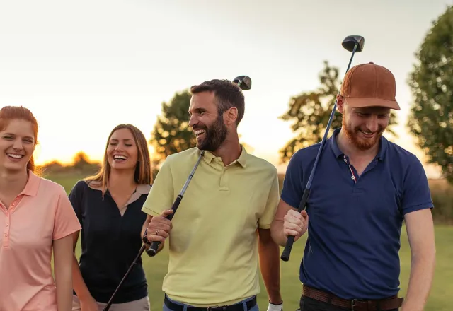 Vier Freunde gehen lachend über den Golfplatz, während im Hintergrund die Sonne untergeht.