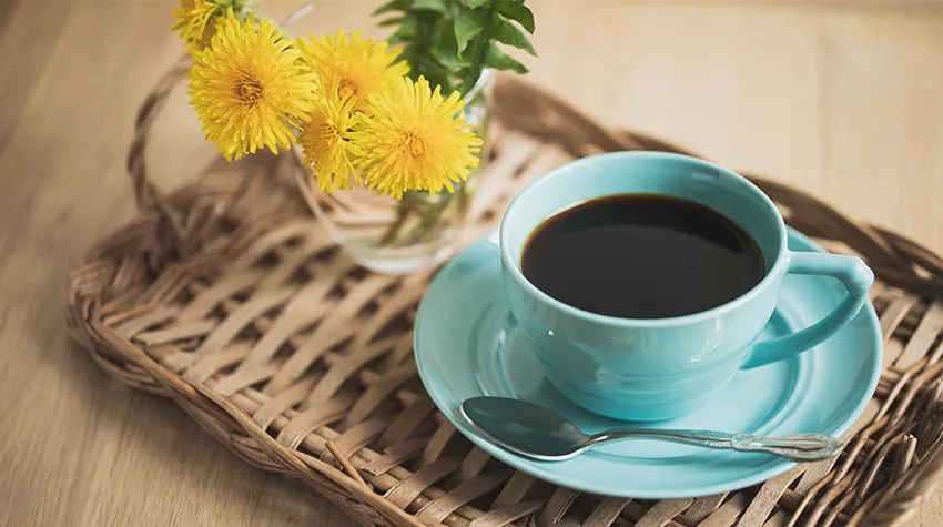 Eine Tasse Kaffee auf einem geflochtenen Tablett. Daneben ein Strauß Löwenzahn.