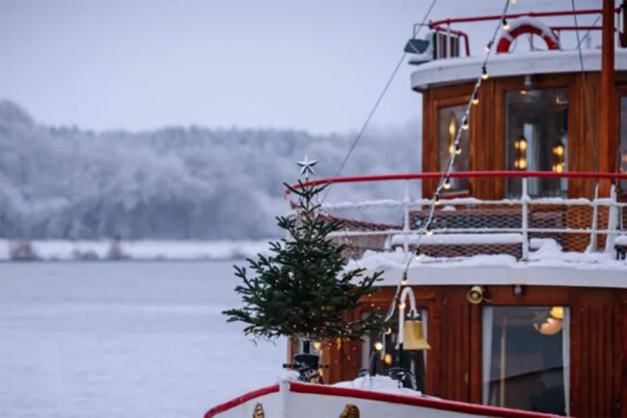 Ein Schiff auf offener See, dessen Bug mit einem Weihnachtsbaum geschmückt ist, festlich umrahmt von leuchtenden Lichterketten.