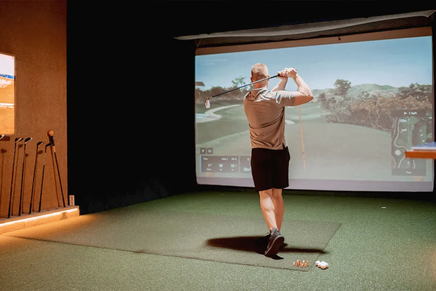 Gast beim Abschlag im Indoor Golf-Simulator an der Ostsee vor virtueller Golfplatzprojektion.
