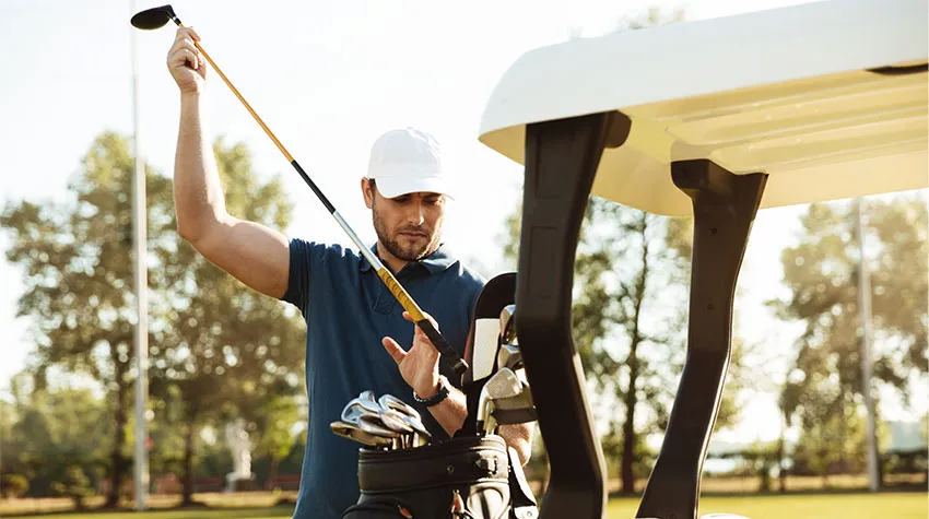 Ein Mann steht am Golfcart und sucht den passenden Schläger aus seiner Golfbag.