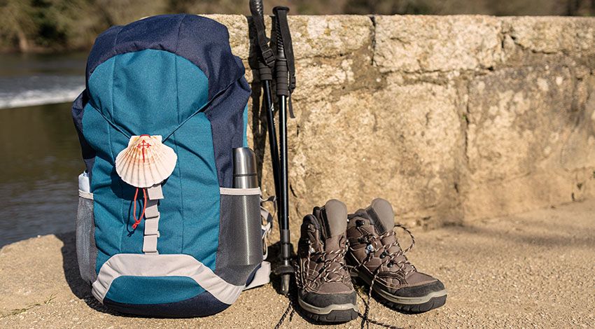 Rucksack mit Muschelnanhänger, daneben Wanderstöcke und Wanderschuhe.