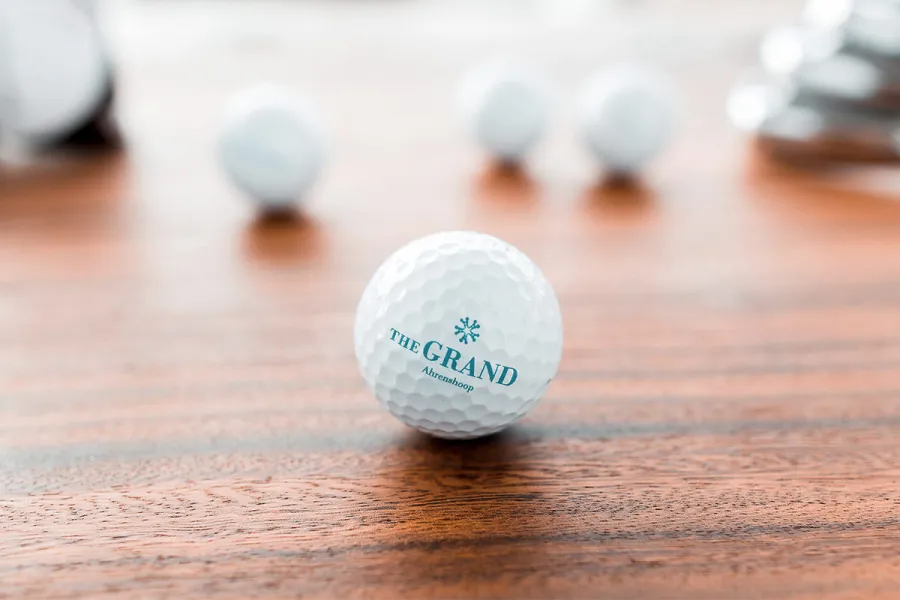 Golfball mit Logo des THE GRAND Ahrenshoop liegt auf Holztisch, weitere Bälle unscharf im Hintergrund.