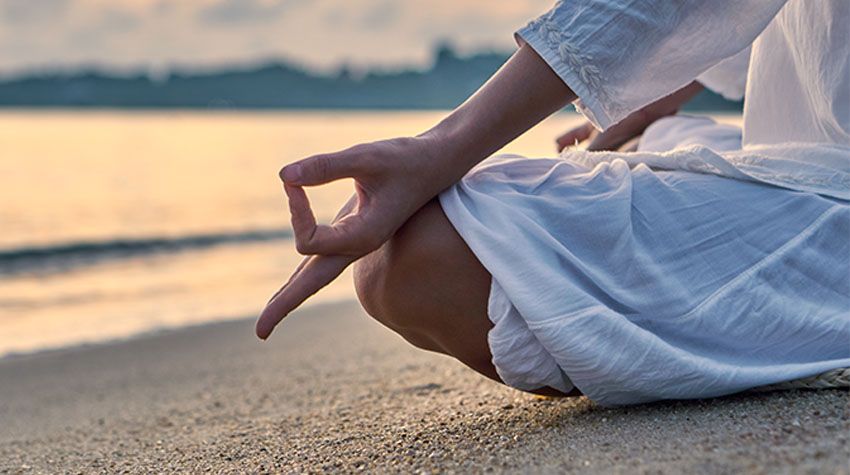 Eine Person sitzt am Strand der Ostsee in Meditationshaltung und genießt den Sonnenuntergang.