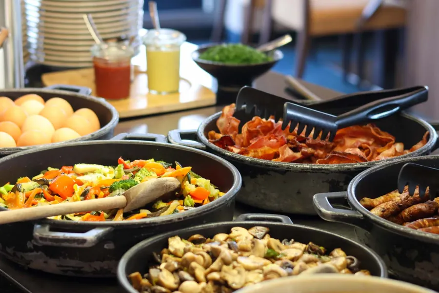 Warmes Frühstücksbuffet an der Ostsee mit Rührei, gebratenem Gemüse, Speck, Würstchen und Pilzen in Pfannen.