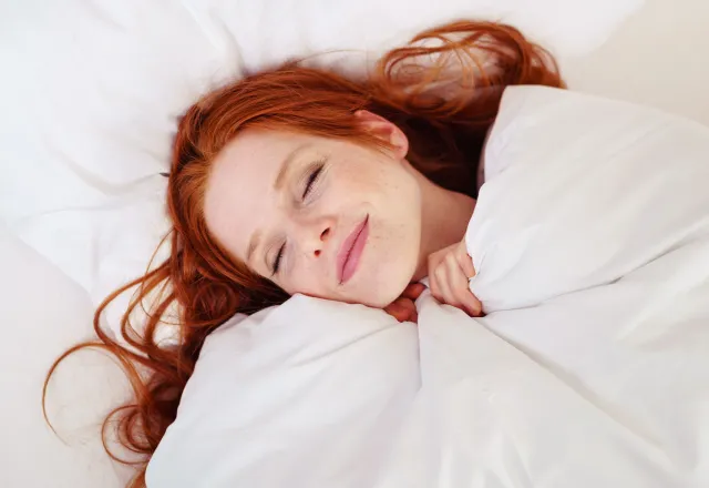 Eine Frau mit roten Haaren liegt gemütlich eingekuschelt im Hotelbett an der Ostsee.