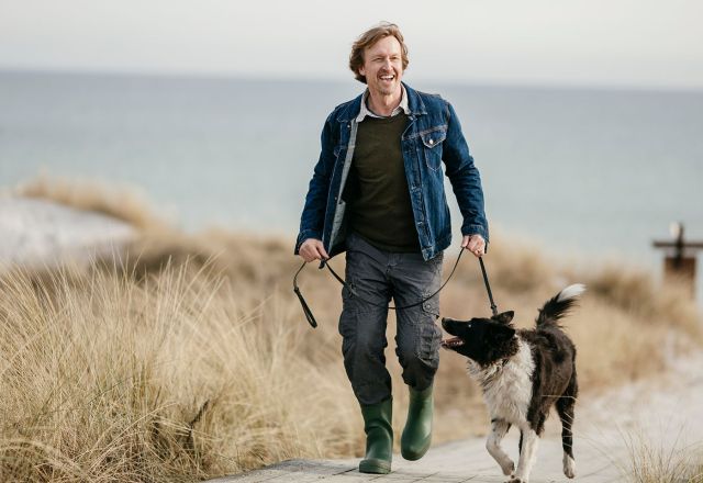 Mann mit Hund an der Leine spaziert entlang eines Strandes. Im Hintergrund Dünen und Wasser.