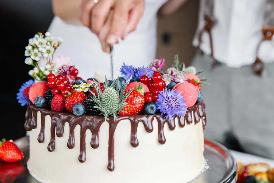 Eine weiße Hochzeitstorte mit Schokoladenverzierung sowie Obst und Blütendekoration wird angeschnitten.