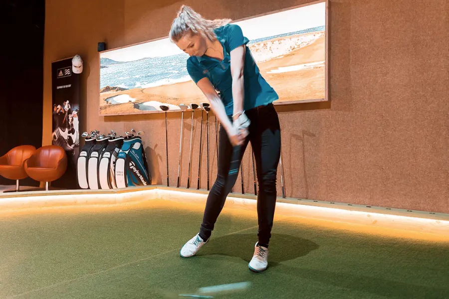 Frau trainiert ihren Golfschwung an der Ostsee auf einer Indoor-Golfanlage mit Schlägern im Hintergrund.
