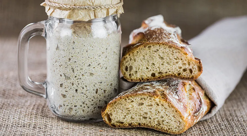 Sauerteig Masse im verschlossenen Glas und daneben zwei Sauerteig Laibe Brot.