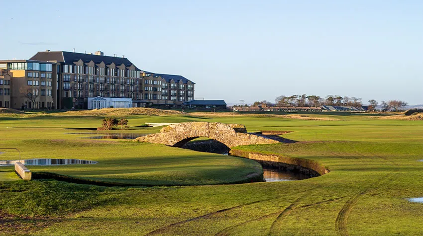 Der Golfplatz Old Course in St Andrews mit Brückenelementen zwischen den Grüns und großen Gebäuden im Hintergrund.