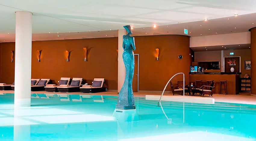 Moderner Innenpoolbereich des THE GRAND Ahrenshoop mit Liegen und einer Frauenstatue im Wasser.