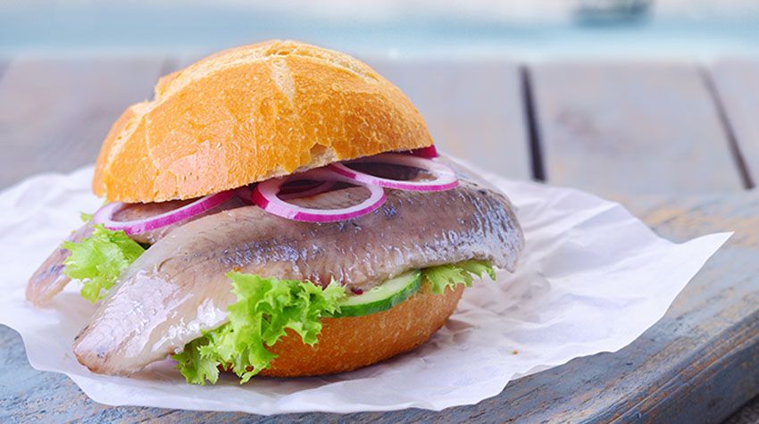 Frisches Fischbrötchen mit Matjes, Salat, Gurke und roten Zwiebeln auf einem Holztisch.