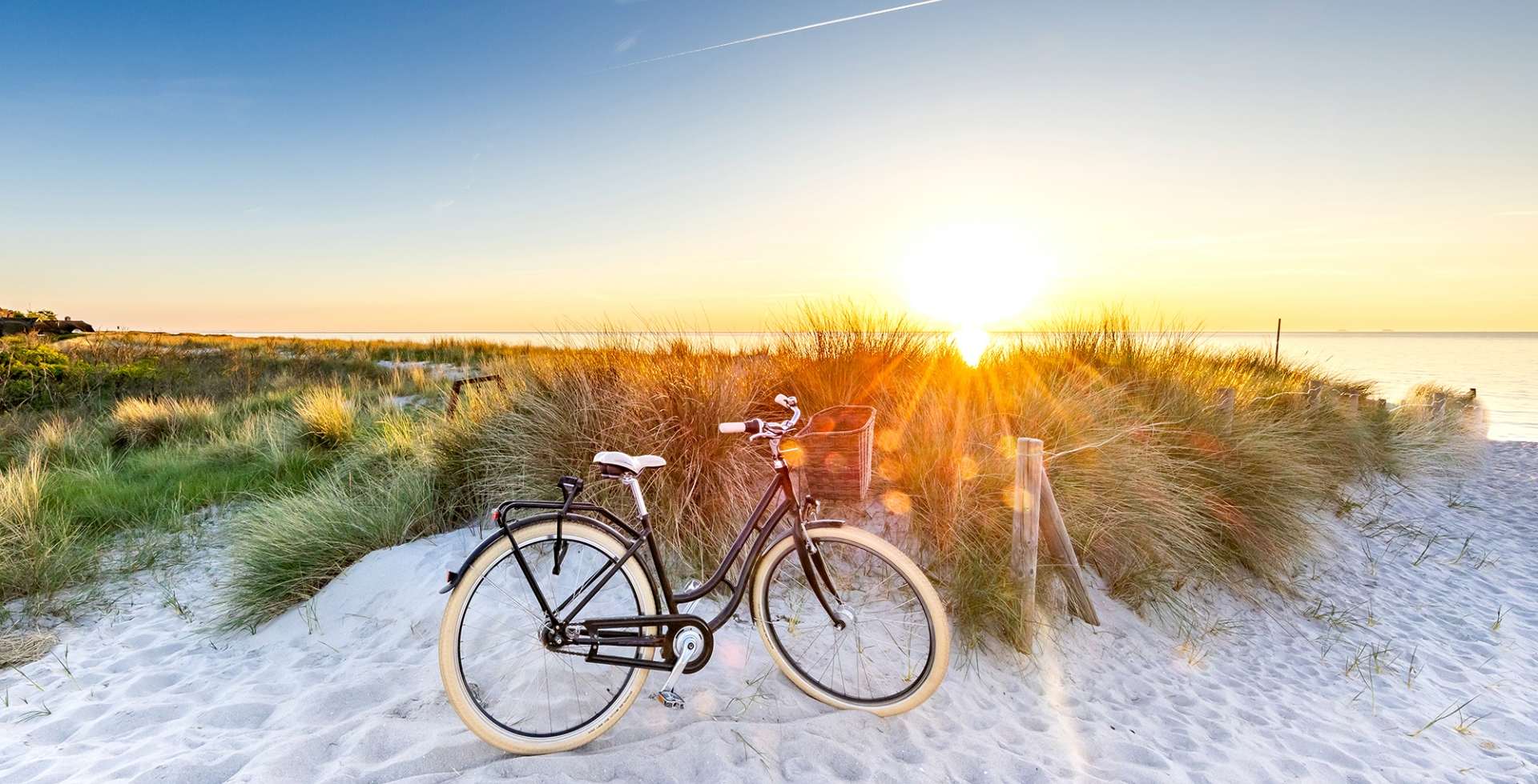 Ein schwarzes Fahrrad steht im Sand vor Dünen am Strand, während die Sonne am Horizont untergeht und warmes Licht über Meer und Gras wirft.