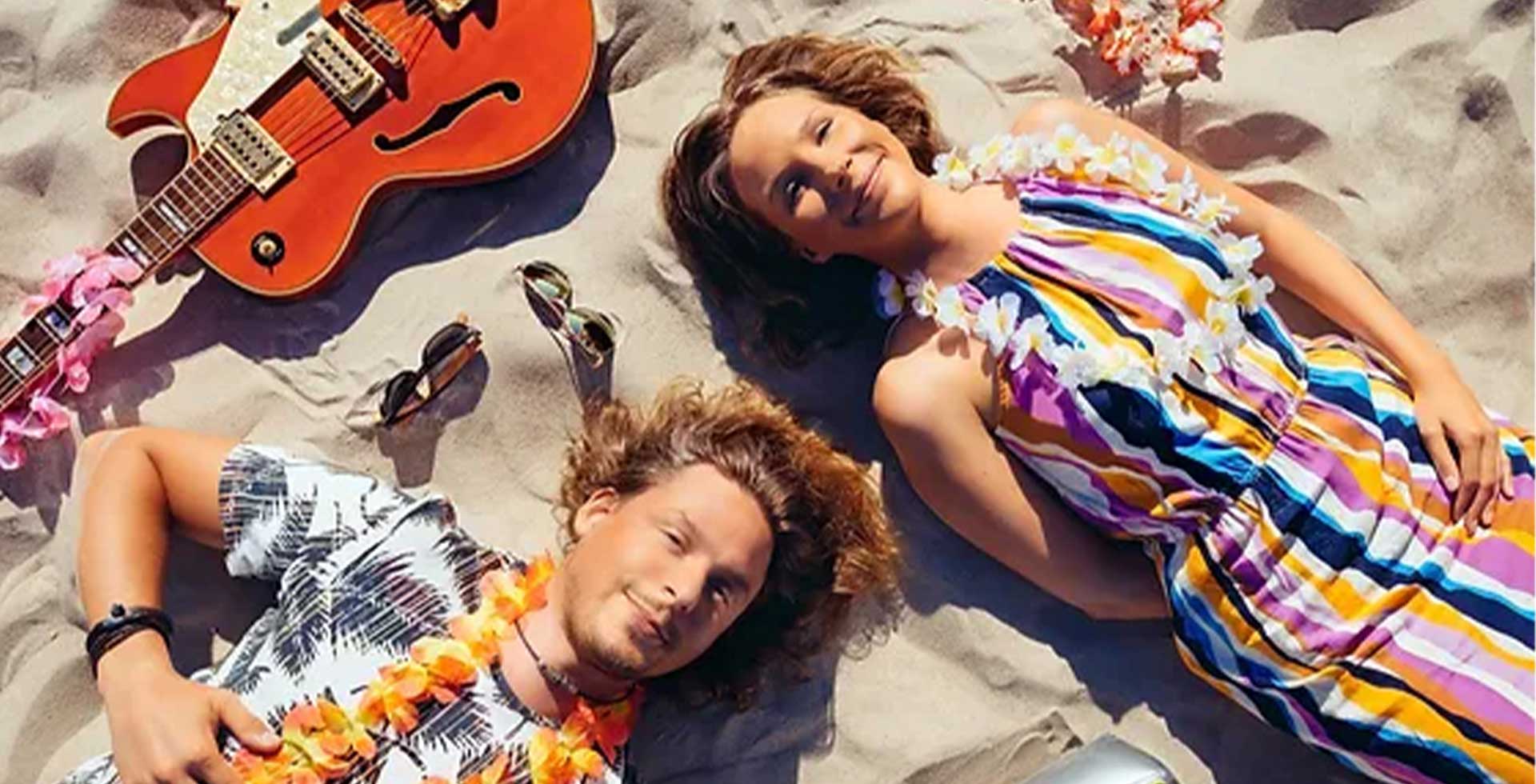 Sommerlich gekleidete Musiker entspannen mit Gitarre am Strand, warmes Licht und lockere Stimmung beim Weihnachtskonzert im THE GRAND.