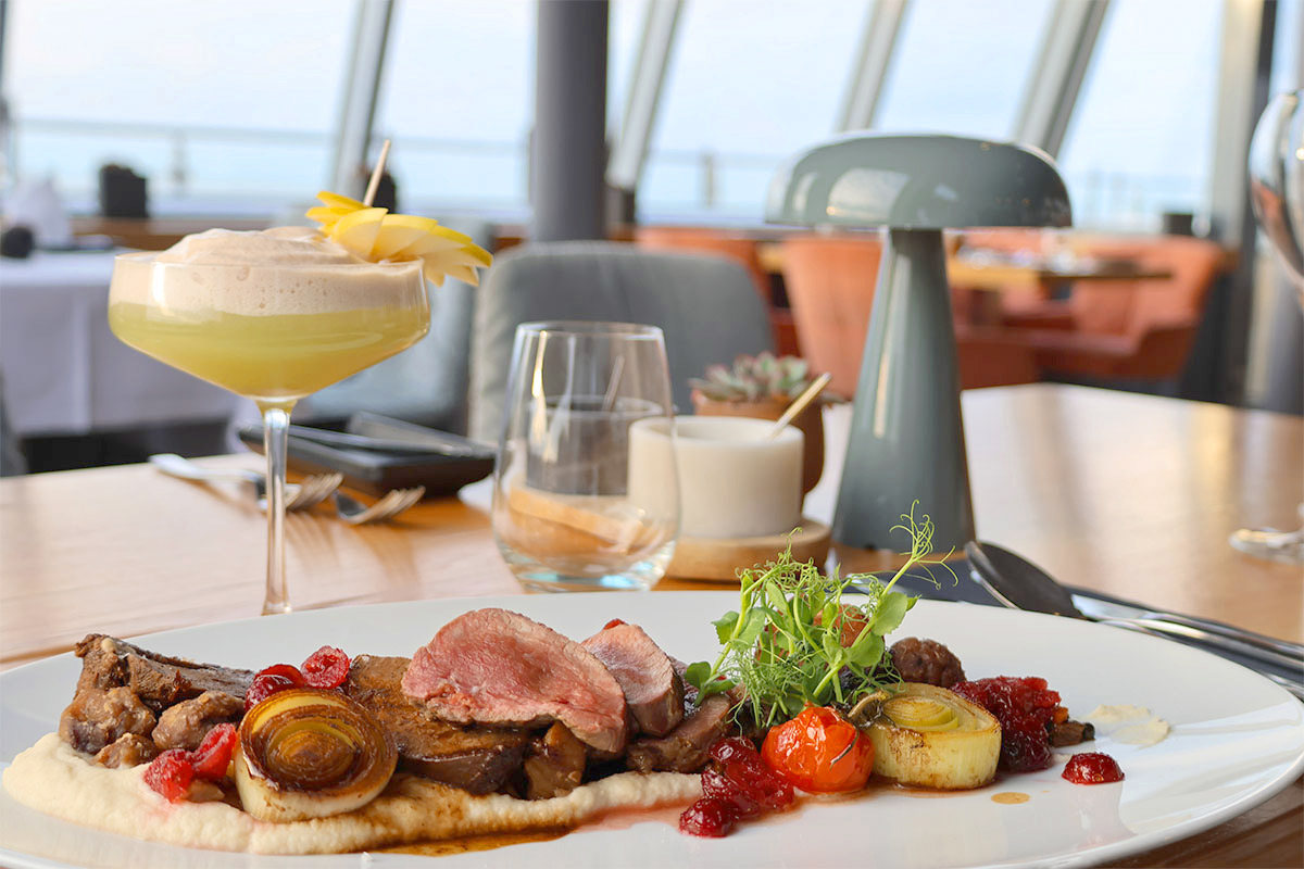 Hauptgericht im Restaurant des THE GRAND Ahrenshoop mit Cocktail und Ostseeblick im Hintergrund.